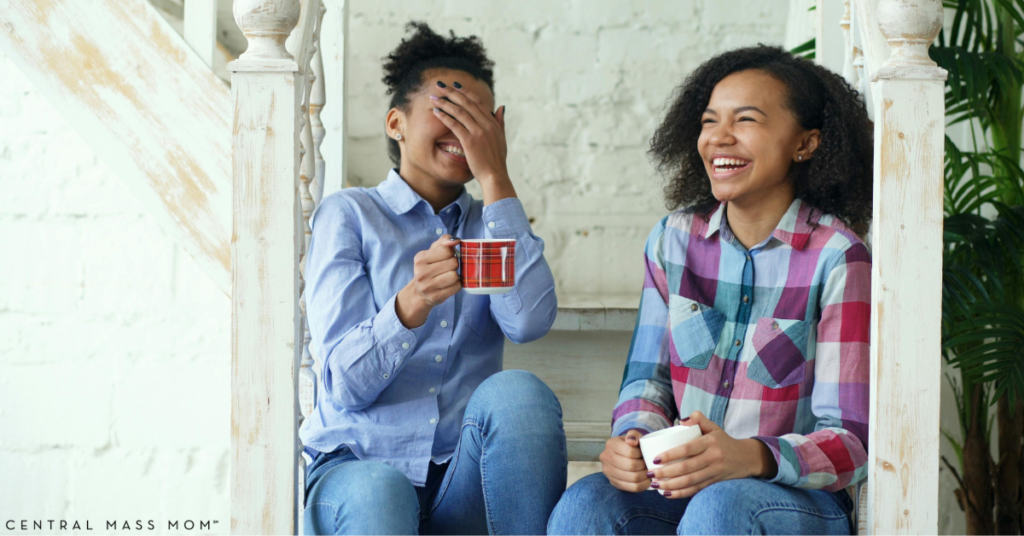 Two women sitting on indoor wooden stairs, laughing heartily and holding coffee mugs. One woman in a blue shirt covers her face while laughing, capturing a relatable moment of shared motherhood stories and "honest" conversation.